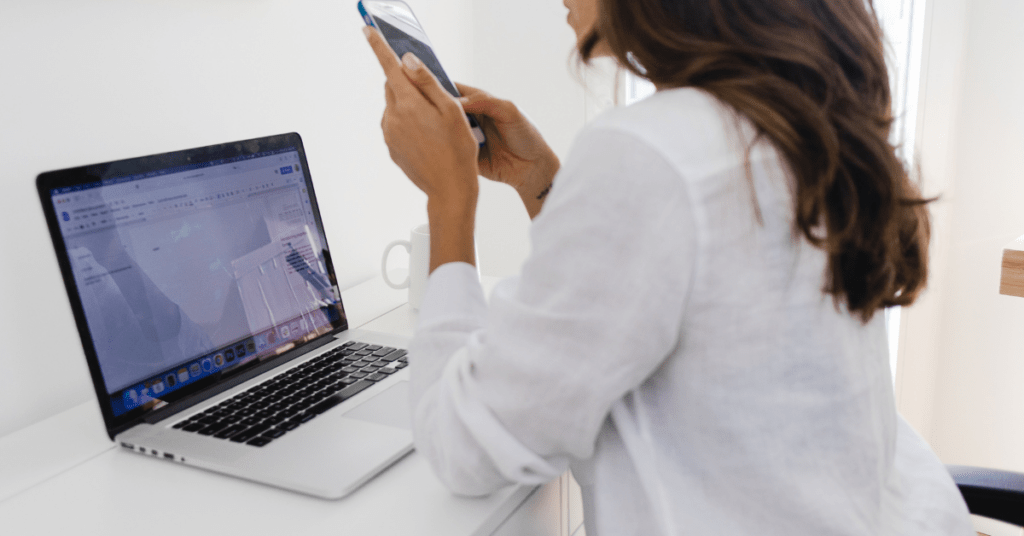 Woman in a white work shirt working at a white metal desk with a silver laptop and using her phone as she struggles with managing digital distractions